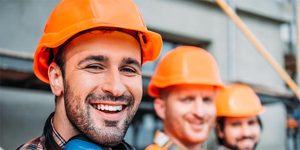Three smiling tradesmen in a line wearing orange safety helmets and vests